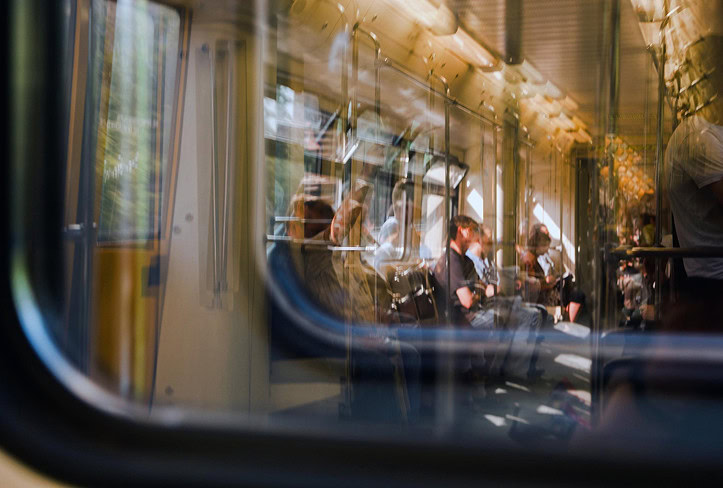 Passengers sit and stand inside a train carriage, seen through a window with reflections and blurred motion, under natural and artificial light.