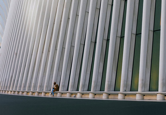 A person sits alone against a large, modern building facade with tall, vertical white panels and narrow windows.
