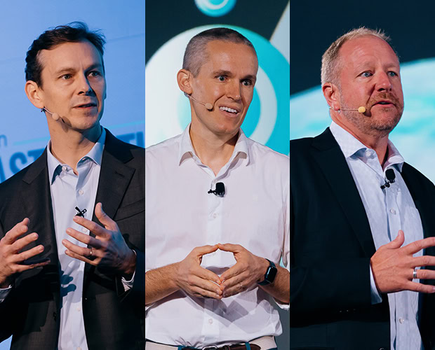Three men, dressed in professional attire, stand separately on stage, each speaking into a microphone while presenting at the registration event.
