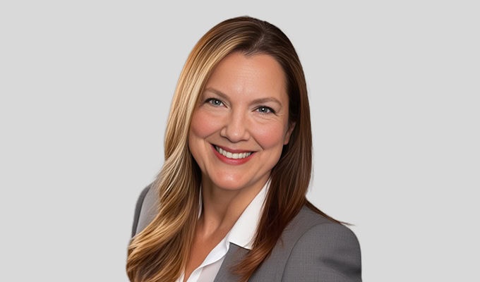 Eine Frau mit langem, glattem, hellbraunem Haar, die einen grauen Blazer und ein weißes Hemd trägt und vor einem schlichten hellgrauen Hintergrund in die Kamera lächelt.