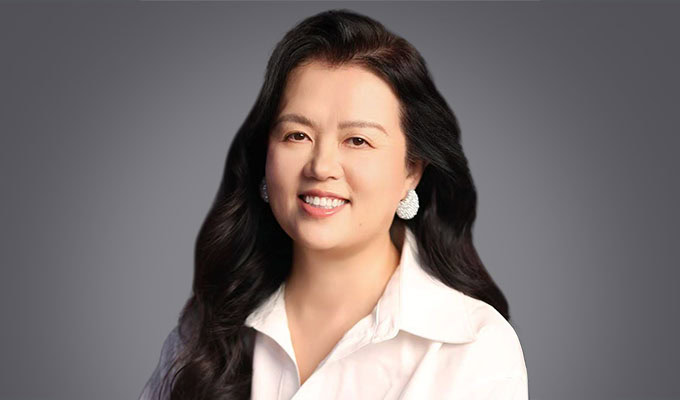 A woman with long black hair, wearing a white collared shirt and earrings, smiles at the camera against a plain gray background.